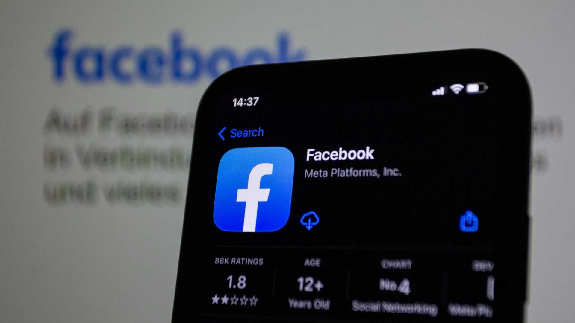 Smartfon wyświetlający ikonę aplikacji Facebook na czarnym tle, w tle widoczny fragment strony Facebooka
